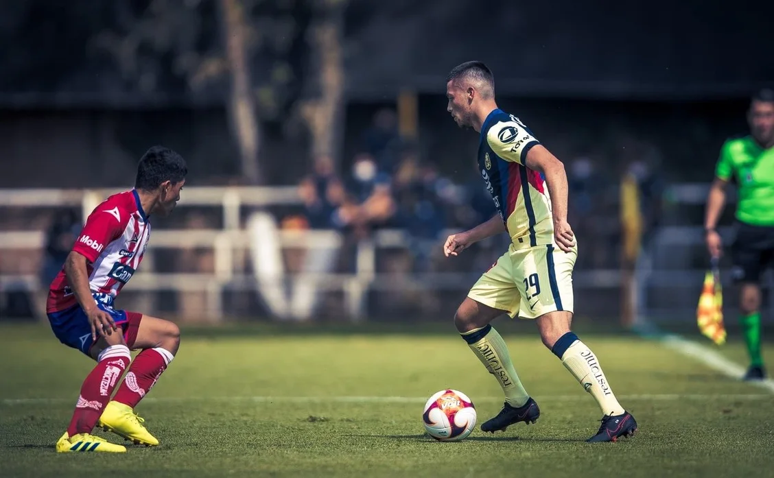 Adrian Goransch, number 29, in Club América's cream and navy kit, facing an opponent in red and white stripes