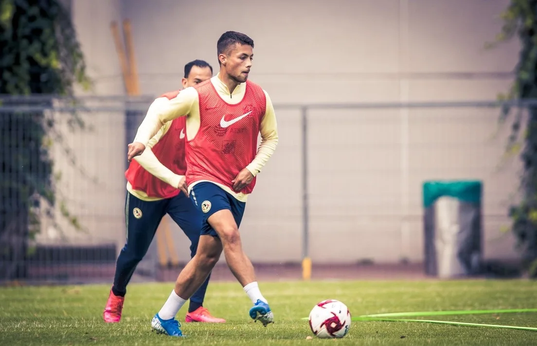 Adrian Goransch striking a ball in a red Club América training bib, a teammate behind him
