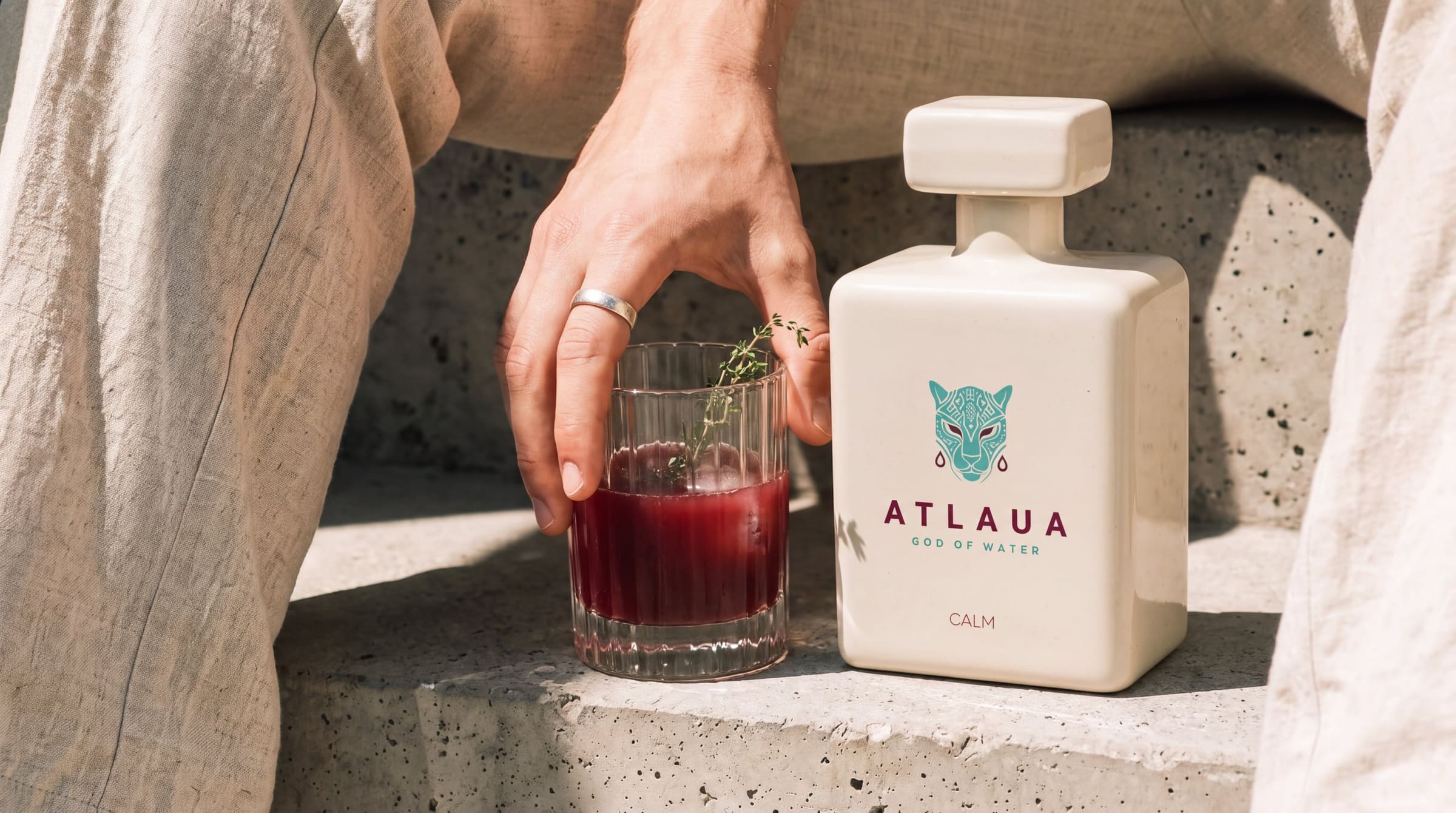 A hand lifting a glass of ATLAUA beside the CALM bottle on sunlit stone steps