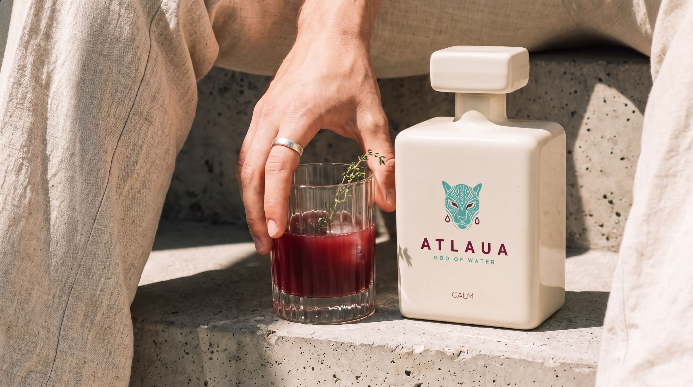 A hand resting on a ribbed glass of berry juice beside a bottle of ATLAUA CALM on sunlit stone steps