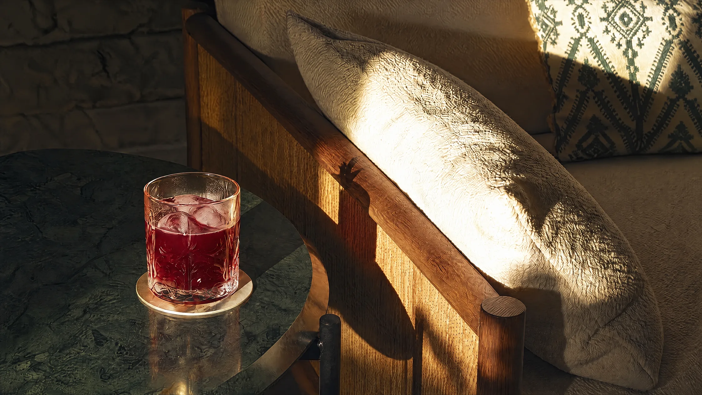 A deep red drink over ice on a green marble table, beside a wooden armchair in late sun